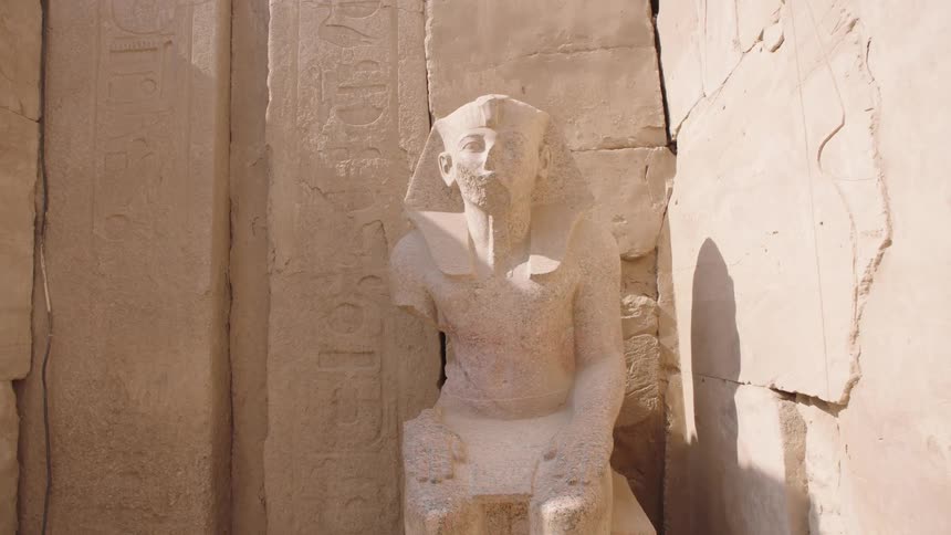 A slow zoom-out shot of a weathered stone statue of an ancient Egyptian pharaoh sitting amongst the ruins of a temple