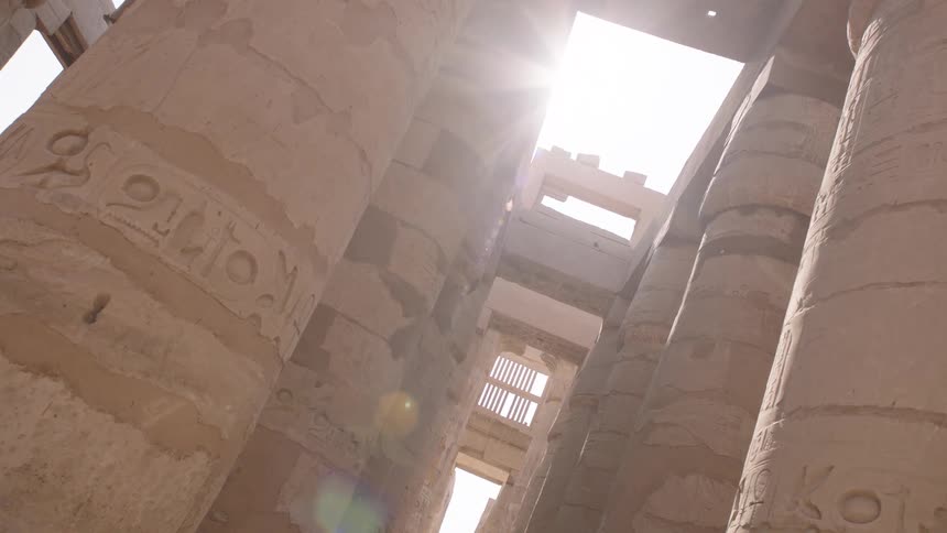 Slow dolly backwards through the massive, sunlit stone columns covered in hieroglyphics at the ancient Karnak Temple in Egypt