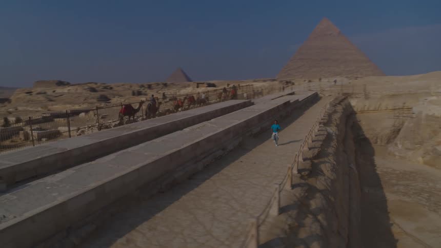 A sweeping pan from the Great Sphinx reveals a man running past tourists on camels towards the magnificent Pyramids of Giza