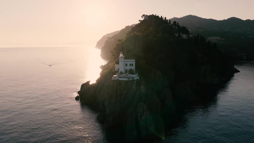 An aerial view reveals a picturesque lighthouse perched atop a dramatic cliffside as the golden sun rises over the calm sea