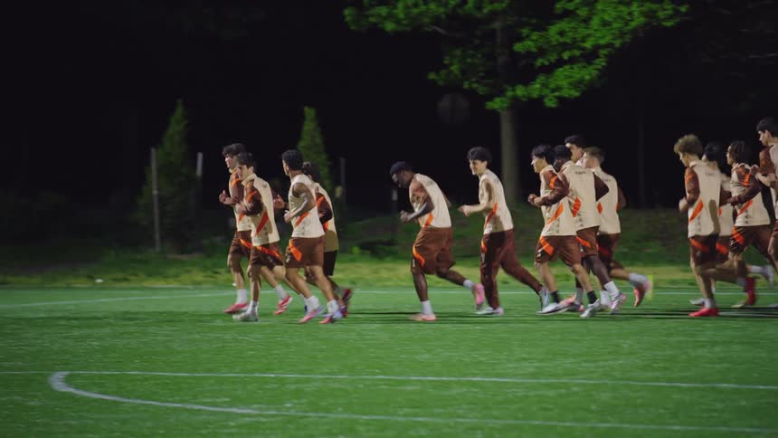 A team of male soccer players jogs in unison across an illuminated field during a training session at night