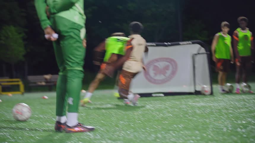 A young men's soccer team participates in aggressive one-on-one dribbling drills and running exercises during a night practice on a turf field
