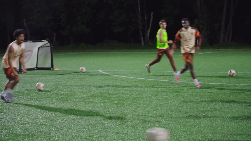 A team of young soccer players practices drills in slow motion on a turf field at night