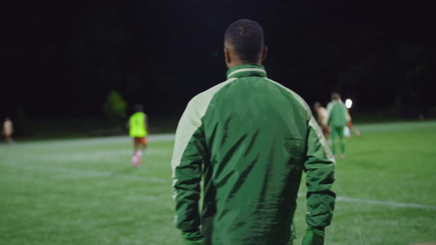 A dedicated soccer coach watches his team from the sidelines during a late-night practice on a floodlit field