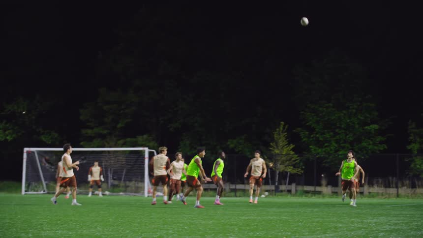 A soccer team practices a header play in slow motion on a lit field at night while their coach watches from the sideline