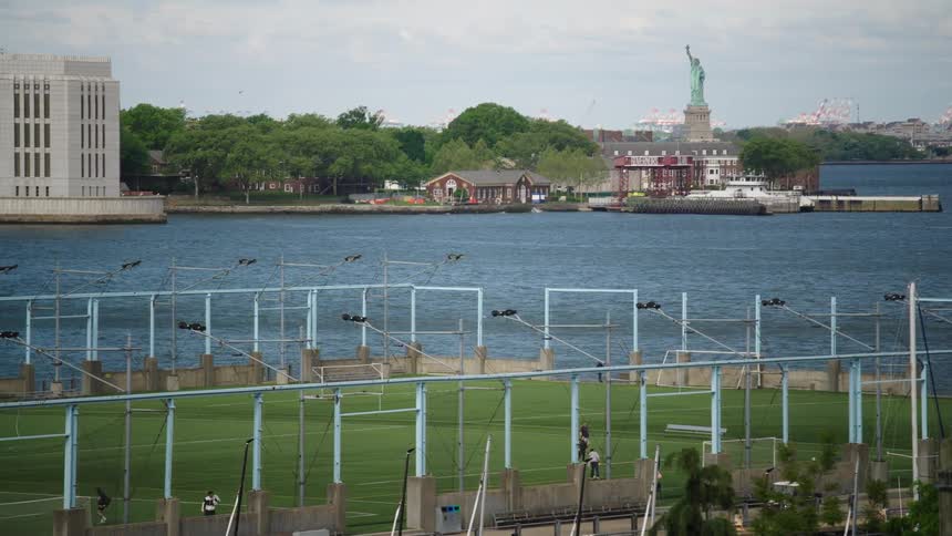 A scenic wide shot of a waterfront sports field in New York City with Ellis Island and the Statue of Liberty visible in the background