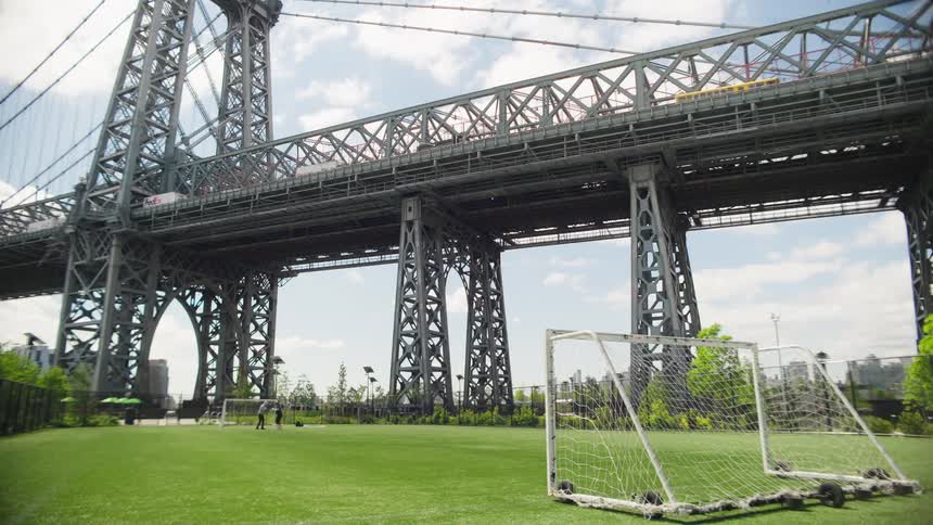 An urban soccer field sits under the Williamsburg Bridge as a subway train crosses above