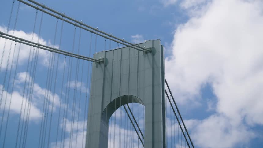 An architectural close-up of a large suspension bridge tower with thick cables against a partly cloudy blue sky