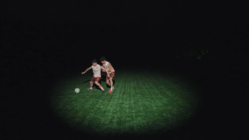 Two male soccer players dribble and defend in slow motion under a spotlight on a dark field at night