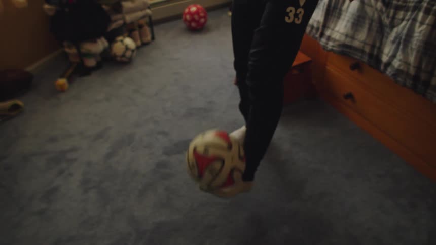 A skilled teenage soccer player expertly juggles a ball with his feet inside his bedroom in slow motion