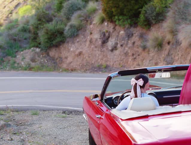 A woman with a pink bow in her hair sits in a vintage red convertible with a surfboard, parked on the side of a scenic country road