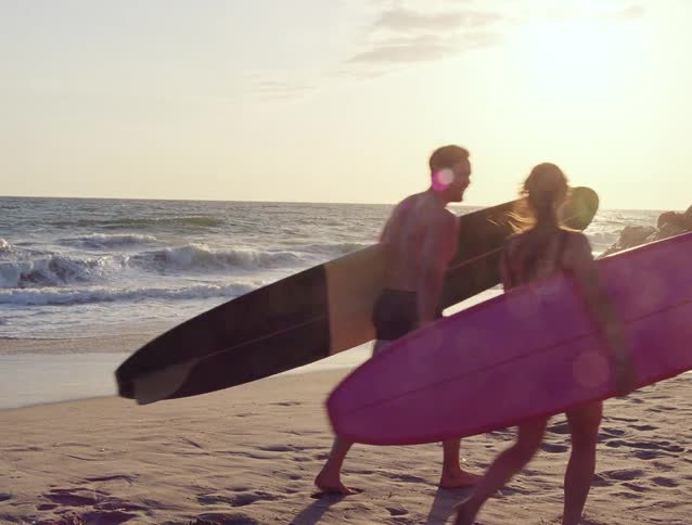 A couple with surfboards walks along the sandy beach shoreline towards the ocean during a warm, golden sunset