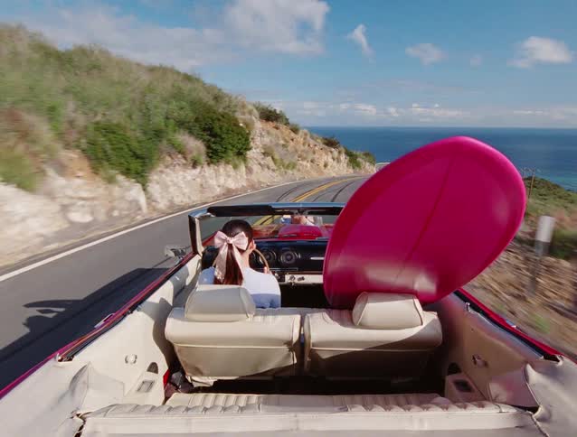 A woman with a pink bow in her hair drives a pink vintage convertible with a matching surfboard down a scenic coastal highway overlooking the ocean