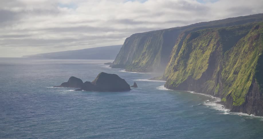 An epic and serene view of the majestic green cliffs of the Pololū Valley coast meeting the Pacific Ocean on a hazy day