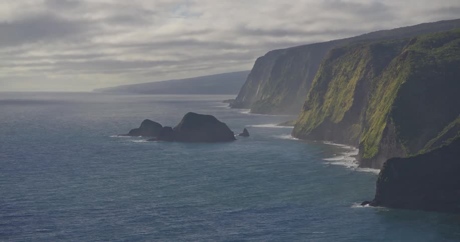 A stunning, wide vista of the dramatic, green-covered cliffs of the Hawaiian coastline meeting the vast, blue ocean under an overcast sky