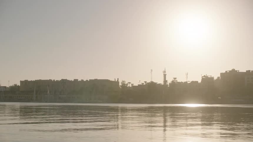 Sunlight glints off the calm waters of the Nile River, revealing the hazy silhouette of a city skyline in Egypt