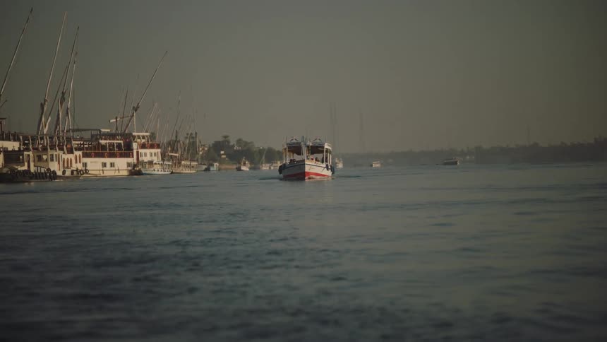 A small tour boat travels along the hazy Nile River in Egypt, passing by a harbor of traditional feluccas