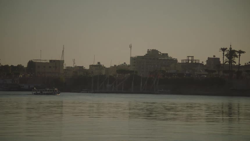 A serene view of the city of Luxor as seen from the Nile River at sunset, with a tour boat sailing past traditional feluccas docked on the shore
