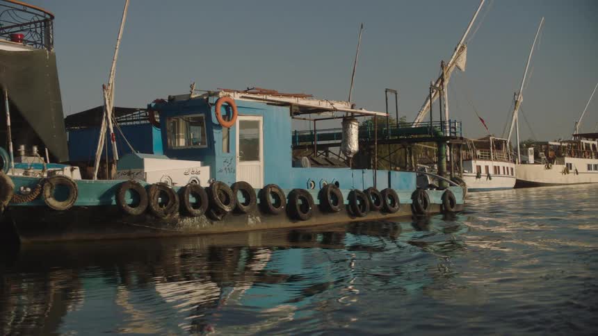 Passing by a blue workboat and other vessels moored at a dock on a sunny day on the Nile River