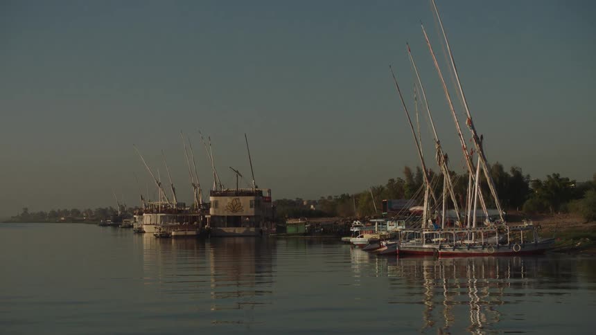 Traditional Egyptian Felucca sailboats and cruise ships are docked along the calm banks of the Nile River at sunrise