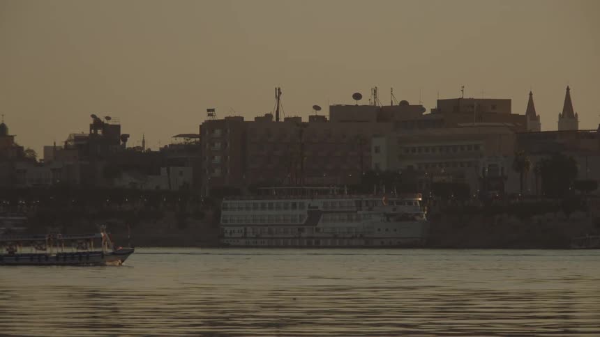 Tour boats travel along a river past a docked cruise ship and a city skyline at sunset