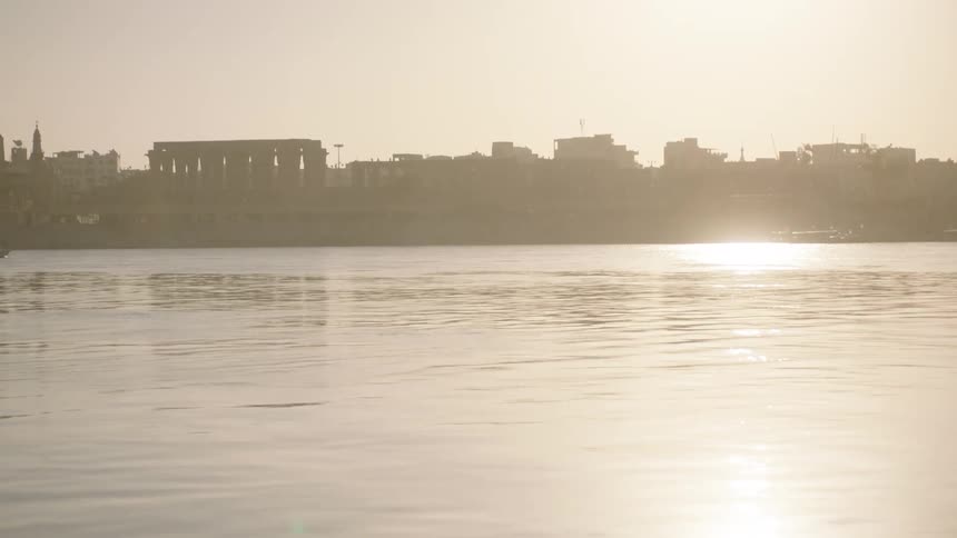Slow tilt up from the sun's reflection on the Nile River to reveal the Luxor Temple silhouetted at sunset