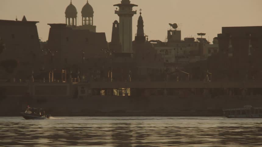 A slow pan across the Nile River shows boats passing by the silhouetted Luxor Temple and city skyline at golden hour