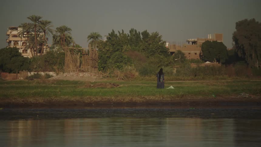 A woman in traditional attire walks along the verdant banks of the Nile River in a rural Egyptian village