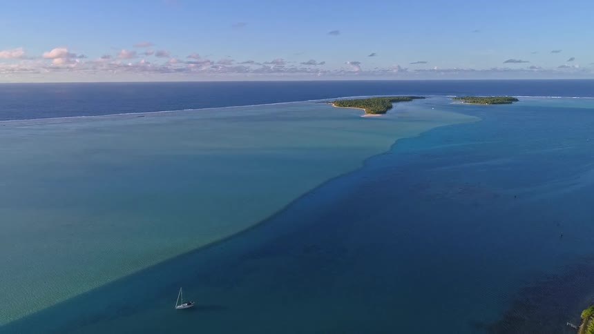 A majestic aerial pullback reveals a sailboat on the breathtaking turquoise and deep blue lagoon of a tropical atoll