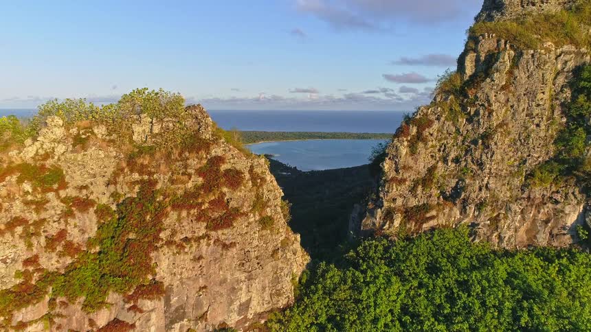 An aerial shot slowly flies through a dramatic mountain pass to reveal a stunning tropical island lagoon at sunrise
