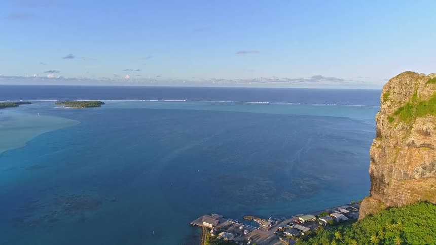 Aerial view slowly reveals a tropical island coastline with a sheer cliff, a small village, and a vast turquoise lagoon