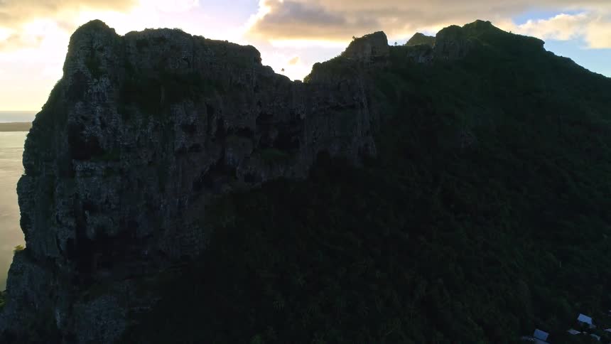 An aerial drone flies over a coastal town toward a towering mountain peak, revealing a brilliant sunburst at sunset on a tropical island
