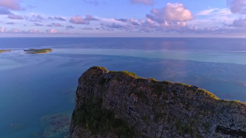 An epic aerial reverse shot reveals a dramatic, lush green mountain peak overlooking a vast tropical blue lagoon and distant islands during a beautiful sunset