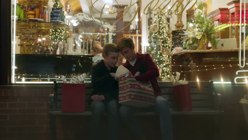 Two boys sitting on bench looking in gift bag in front of shop at night