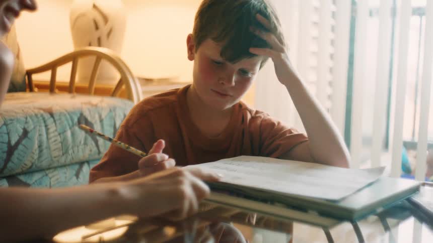 A young boy smiles as he diligently works on his homework at a glass table with a parent nearby