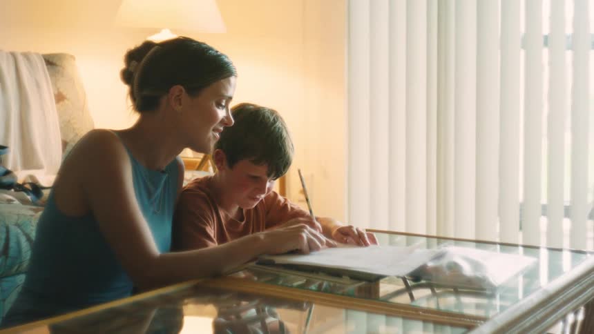 A loving mother watches her son as he happily draws in a warmly lit living room