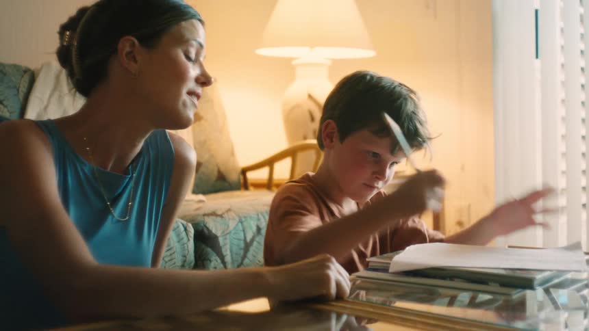 A loving mother smiles and helps her young son with his homework at a glass table in their warmly lit home
