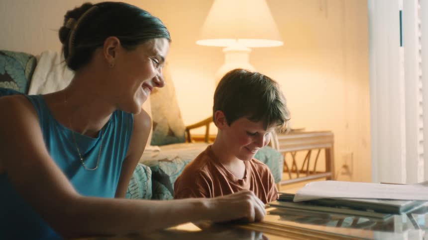 A loving mother smiles and helps her young son with his homework at a glass table in their warmly lit home