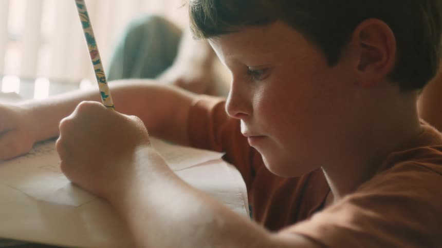 A close up shot of a young boy smiling as he writes with a pencil