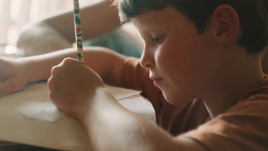 A close up shot of a young boy smiling as he writes with a pencil