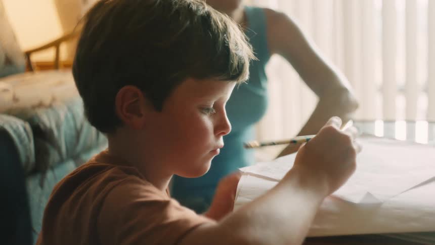 A young boy playfully balances his pencil on his nose before getting back to his schoolwork at a table indoors