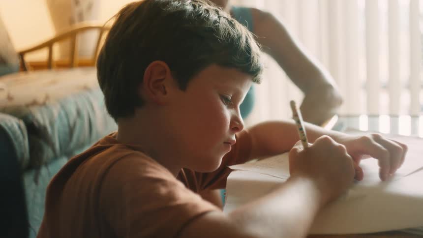 A young boy playfully balances his pencil on his nose before getting back to his schoolwork at a table indoors