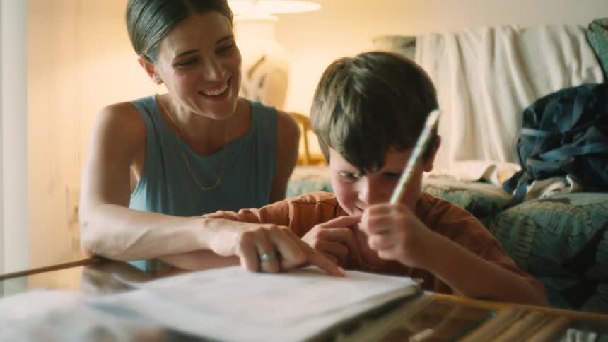 A caring mother proudly watches and helps her young son as he concentrates on his homework at the table