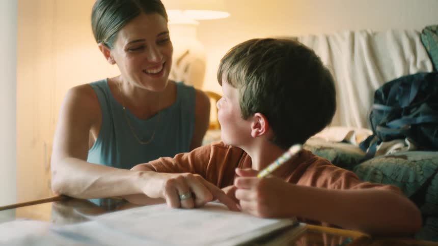 A caring mother proudly watches and helps her young son as he concentrates on his homework at the table