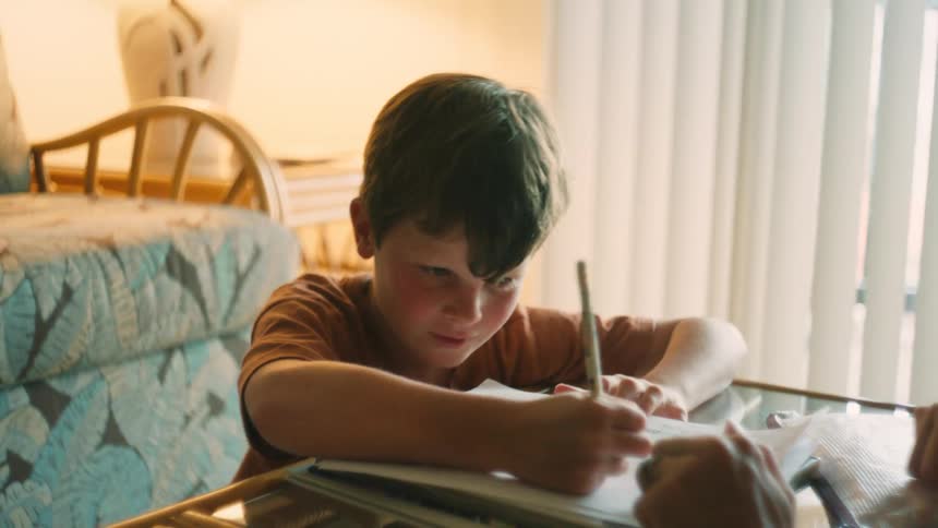 A close-up of a smiling young boy sitting at a table doing his homework in a sunlit room