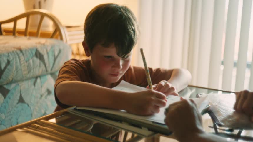 A close-up of a smiling young boy sitting at a table doing his homework in a sunlit room