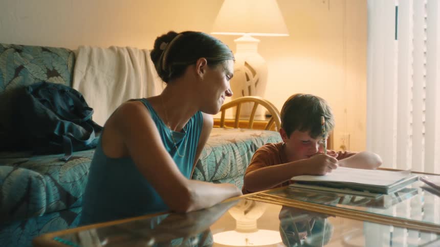 A smiling mother watches and helps her young son as he concentrates on drawing in a notebook at a coffee table in a warmly lit room