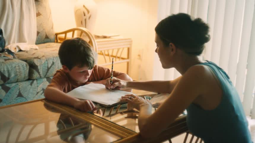 A mother helps her young son with his homework, guiding his hand as he writes with a pencil at a table in their sunlit home