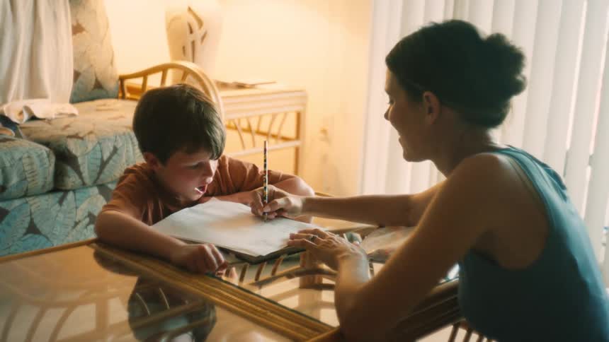 A mother helps her young son with his homework, guiding his hand as he writes with a pencil at a table in their sunlit home