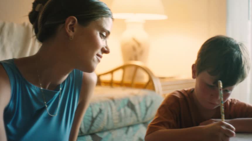 A caring mother smiles lovingly while watching her young son concentrate on his homework at a table in a cozy room